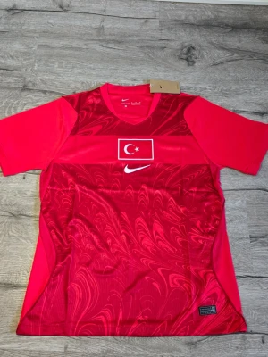 Turkiet matchtröja 2026 - Snygg röd matchtröja från Nike med Turkiets flagga på bröstet och vit Nike-logga. Tröjan har korta ärmar, rund hals och ett mönstrat tyg framtill. På ryggen står det 'Türkiye' tryckt i vitt. Perfekt för dig som vill stötta landslaget.
