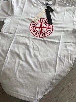 Vit Stone Island t-shirt med rött tryck - Säljer en vit t-shirt från Stone Island med stort rött broderat logotyptryck på bröstet. Klassisk rund hals och korta ärmar. Tillverkad i mjuk bomull för en skön känsla. Perfekt för dig som gillar streetwear och vill sticka ut.
