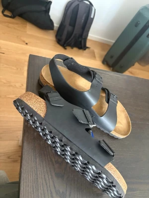 Svarta sandaler med remmar och spännen - Säljer ett par klassiska svarta Birkenstock sandaler med två justerbara remmar och spännen. Fotbädden är i kork och mocka för extra komfort och sulan har ett greppvänligt mönster. Perfekta för dig som gillar stilrena och bekväma skor.Helt oanvända storlek 44.