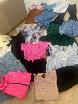 Klädpaket med toppar, klänningar & shorts - Säljer ett klädpaket med flera plagg: rosa sportig t-shirt, blommig topp, grön klänning med knappar, jeansshorts, rosa hoodie, spetsdetaljerad topp, blå skjorta och fler. Färgglada och somriga plagg i olika stilar och material, perfekt för att mixa och matcha. Passar 11 till 12 år 