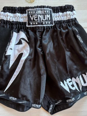 Svarta Muay Thai shorts från Venum - Snygga svarta Muay Thai shorts från Venum med vitt tryck och logga på benet och midjan. Shortsen har bred resår i midjan och meshpaneler på sidorna för extra ventilation. Tillverkade i glansigt, slitstarkt material som passar perfekt för kampsportsträning.
