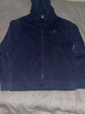 Mörkblå hoodie från Polo Ralph Lauren - Snygg mörkblå hoodie med dragkedja från Polo Ralph Lauren. Klassisk röd logga på bröstet, huva och kängurufickor framtill. Tillverkad i mjukt bomullsmaterial som känns skönt mot huden. Perfekt för en chill och avslappnad stil.