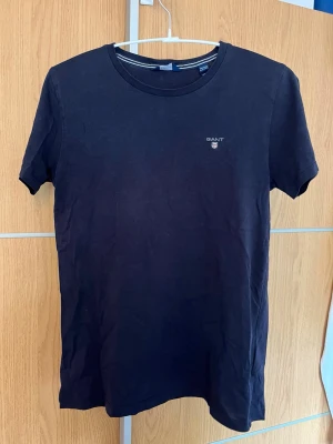 Mörkblå t-shirt från GANT - Snygg mörkblå t-shirt från GANT med liten logga på bröstet. Klassisk rund halsringning och korta ärmar. Tillverkad i mjuk bomull som känns skön mot huden. Perfekt till jeans eller shorts för en clean och enkel stil.