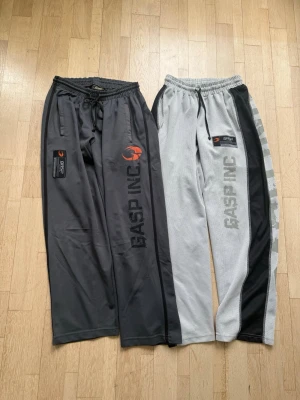 Två par mjukisbyxor från GASP INC - Säljer två par mjukisbyxor från GASP INC. Ena paret är mörkgrått med svart och orange logga, det andra är ljusgrått med svarta detaljer och tryck längs benet. Båda har dragsko i midjan och sidofickor. Perfekta för gymmet eller chill.