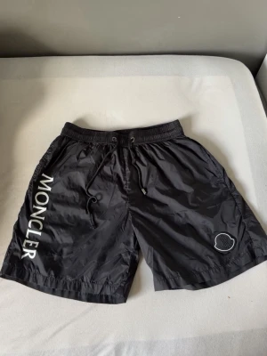 Svarta Moncler shorts med snörning - Snygga svarta shorts från Moncler med vit logga längs benet och broderad patch på ena benet. Shortsen har elastisk midja med snörning och är tillverkade i ett lätt, glansigt syntetmaterial. Perfekta för sommaren och riktigt stilrena.