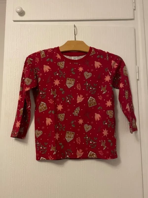 Röd långärmad jul-tshirt Lindex barn - Röd långärmad t-shirt från Lindex med juliga motiv som pepparkakshjärtan, stjärnor, renar och små hus. Perfekt för vinterns mysiga dagar. Tillverkad i mjuk bomull för skön komfort.