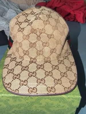 Beige keps från Gucci med GG-mönster - Säljer en beige keps från Gucci med det klassiska GG-monogrammet i brunt över hela kepsen. På sidan finns en grön och röd rand som signaturdetalj. Kepsen har böjd skärm och är tillverkad i canvasmaterial. Perfekt för dig som vill ha en lyxig och trendig accessoar.