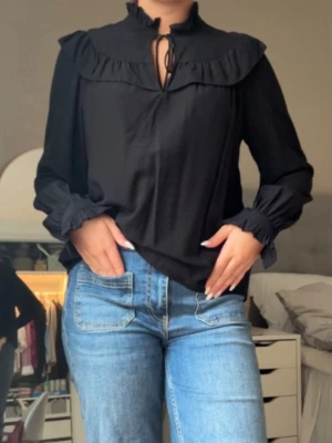 Svart blus med volang och knyt - Stilren svart blus med volangdetaljer längs axlar och hals, samt knytband vid den höga kragen. Blusen har långa ärmar med resår och volang vid ärmslut.