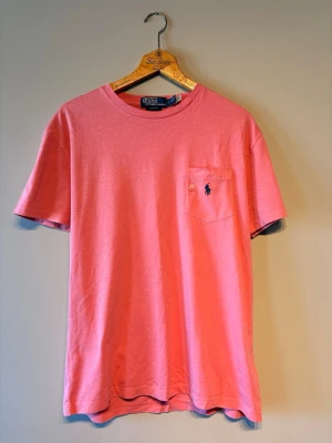 Rosa t-shirt från Polo Ralph Lauren - Snygg rosa t-shirt från Polo Ralph Lauren med klassisk passform. T-shirten har korta ärmar, rund halsringning och en bröstficka med den ikoniska broderade loggan i marinblått. Tillverkad i mjuk bomull för skön känsla. Perfekt skick aldrig använd.