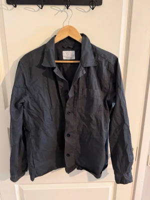 Mörkgrå overshirt från Form&Thread - Stilren mörkgrå overshirt från Form&Thread med klassisk krage, knappstängning och bröstficka. Tillverkad i ett tunt, mjukt bomullstyg som ger en avslappnad look. Perfekt att bära över en t-shirt för en enkel men snygg outfit.