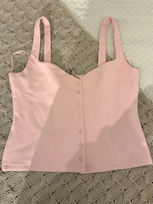 Ljusrosa korsett-topp med knappar - Supersöt ljusrosa korsett-topp med breda axelband och knappar framtill. Toppen har en figurnära passform och är perfekt för att skapa en feminin look. Tillverkad i ett mjukt och stretchigt material som känns bekvämt mot huden. Toppen är en storlek L men jag skulle säga att den är mindre i storleken, den är mer åt en storlek M än L. 