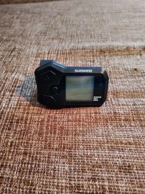 Shimano SC-E5000 cykeldator - En svart cykeldator från Shimano, modell SC-E5000. Den har en digital display och flera knappar för navigering. Tillverkad i plast och designad för att monteras på elcykelstyre. Perfekt för att hålla koll på hastighet, distans och batterinivå.