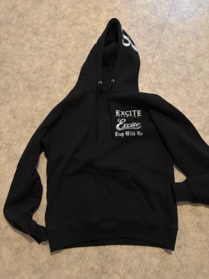 Svart hoodie med tryck och huva - Svart hoodie med stor huva och dragsnören. Tryck i vitt på bröstet med texten 'EXCITE Excite Trap With Us'. Stor magficka framtill och ribbade muddar vid ärmar och nederkant. Perfekt för en avslappnad streetwear-look.