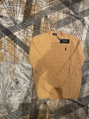 Beige kabelstickad tröja Polo Ralph Lauren - Kabelstickad tröja i beige från Polo Ralph Lauren med rund halsringning och broderad logga på bröstet. Tröjan har ribbstickade muddar och klassisk passform. Perfekt för dig som gillar stilrena och tidlösa plagg.