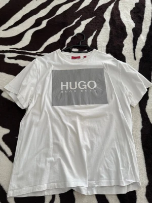 Vit HUGO BOSS t-shirt med logga - Vit t-shirt från HUGO BOSS med stor grå logga på bröstet. Tillverkad i 100% ekologisk bomull, klassisk passform och rund halsringning. Perfekt för en stilren och enkel look.