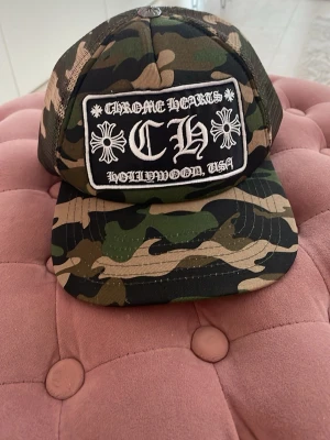 Camo keps Chrome Hearts - Säljer en camo keps från Chrome Hearts med stort svartvitt patch framtill och broderad text. Kepsen har klassiskt böjd skärm och mesh på sidorna för extra ventilation. Justerbar baktill för perfekt passform.