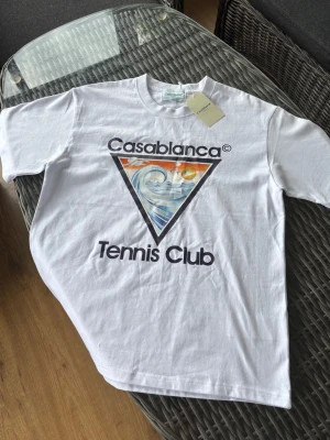Vit Casablanca Tennis Club t-shirt - Säljer en vit t-shirt från Casablanca med stort färgglatt tryck på bröstet där det står 'Casablanca Tennis Club' och en våg i en triangel. Klassisk passform och rund hals. Perfekt för dig som gillar streetwear och grafiska prints.