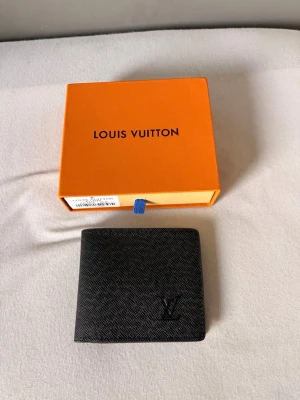 Svart Louis Vuitton plånbok i läder - Snygg svart plånbok från Louis Vuitton i exklusivt präglat läder. Klassisk vikbar modell med flera kortfack, fönster för ID och diskret LV-logga på framsidan. Perfekt för dig som vill ha stilren och lyxig accessoar.