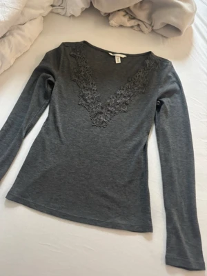 Mörkgrå långärmad topp med spets - Mörkgrå långärmad topp från H&M med v-ringning dekorerad med spetsdetaljer framtill. Tunn och mjuk i materialet, perfekt för lager på lager eller som den är. Enkel men ändå med en snygg twist tack vare spetsen.