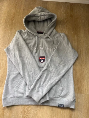 Grå hoodie med broderad logga - Snygg grå hoodie med broderad text 'MARINE' och en patch med marinblå och röda detaljer på bröstet. Huva med dragsko och magficka framtill. Ribbstickade muddar vid ärmslut och nederkant. Perfekt för en avslappnad stil.