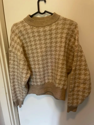 Beige stickad tröja med hundtandsmönster - Säljer en beige stickad tröja med klassiskt hundtandsmönster i ljusbeige och sandfärg. Tröjan har bred ribbad mudd i hals, ärmslut och nederkant samt en avslappnad passform med droppade axlar. Perfekt för dig som gillar retrovibbar och mjuka material.