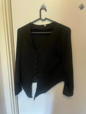 Svart asymmetrisk blus med knappar - Snygg svart blus med v-ringning och knappar framtill. Blusen har lång ärm och en unik asymmetrisk nederkant som ger en cool look. Perfekt för dig som vill ha något stilrent men ändå lite edgy. Materialet känns lätt och bekvämt.