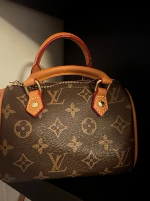monogram handväska - Säljer en klassisk brun handväska med det ikoniska monogrammönstret i beige och guldiga metalldetaljer. Väskan har rundade handtag i läder och dragkedja upptill. Perfekt accessoar för dig som gillar lyxig och tidlös design.