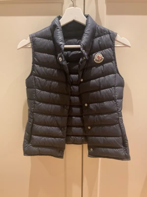 Svart dunväst från Moncler - Snygg svart dunväst från Moncler med hög krage och tryckknappar framtill. Västen har ett klassiskt Moncler-märke på bröstet och är quiltad för extra värme. Perfekt att slänga över en hoodie eller tröja för en trendig lager-på-lager-look.