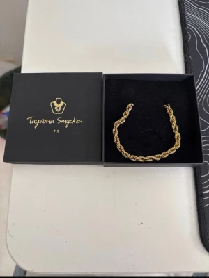 Flätat halsband TAYRONA SMYCKEN - Snyggt flätat halsband i guldfärg från Tayrona Smycken. Halsebanet har en tvinnad design som ger en exklusiv känsla och passar perfekt till din stil. Levereras i original ask.
