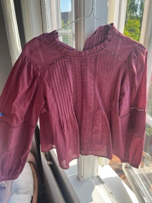 Vinröd blus med spetsdetaljer - Säljer en vinröd blus med långärmad design och fina spetsdetaljer längs ärmar och framtill. Blusen har rundad halsringning med volangkant och dekorativa veck framtill. Tillverkad i ett tunt, luftigt tyg som ger en romantisk känsla.