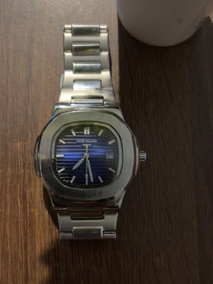 Klocka  - Stilren klocka med blå urtavla och silverfärgad länk i metall. Klockan har datumvisning och klassisk design med rektangulär boett och tydliga indexmarkeringar. Perfekt accessoar för dig som gillar exklusiva detaljer.