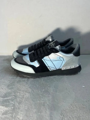 Valentino Garavani blå sneakers - Snygga sneakers från Valentino Garavani i blått, svart och grått med coolt kamouflagemönster. Skorna har svarta mockadetaljer, vita och ljusblå partier samt nitar på hälen. Tjock sula och klassisk snörning. Perfekta för dig som vill sticka ut.