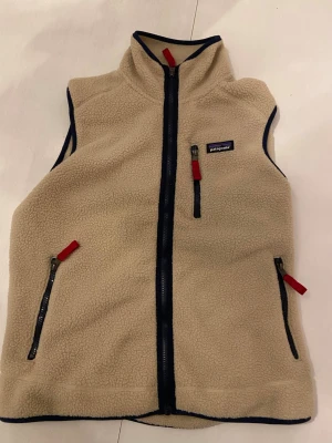 Beige fleeceväst från Patagonia - Mysig beige fleeceväst från Patagonia med marinblå detaljer och röda dragkedjor. Västen har hög krage, tre fickor med dragkedja och ett klassiskt Patagonia-märke på bröstet. Perfekt för lager-på-lager och outdoor-stil.