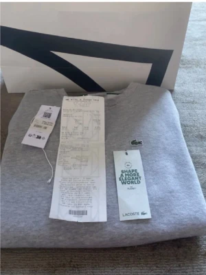 Grå hoodie från Lacoste - Snygg grå hoodie från Lacoste med den klassiska gröna krokodilloggan broderad på bröstet. Hoodien har ribbade muddar vid ärmslut och nederkant samt en mjuk, bekväm bomullskvalitet. Perfekt för en clean och stilren look.