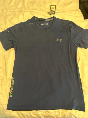 Blå tränings-tshirt från Under Armour - Snygg blå tränings-tshirt från Under Armour med rund hals och korta ärmar. Logga på bröstet och färgglad text längs sidan. Tillverkad i ett lätt och snabbtorkande funktionsmaterial, perfekt för löpning eller gym.