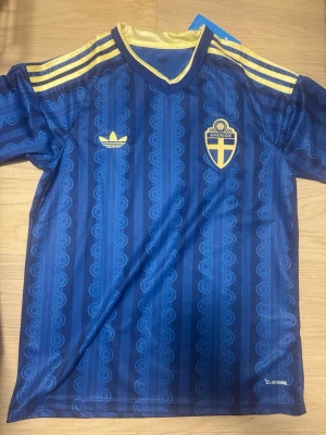 Blå Sverige fotbollströja Adidas - Säljer en blå fotbollströja från Adidas med Sveriges landslagsemblem på bröstet. Tröjan har gula detaljer på axlarna, V-ringad krage med gul kant och ett retroinspirerat mönster. Tillverkad i lätt och ventilerande material (Climacool). Perfekt för match eller träning.