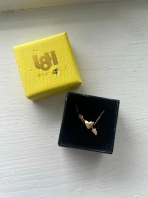 Guldigt halsband l8hasselblad - Säljer ett guldigt halsband med ett hjärthänge och pil, levereras i original gul ask. Kedjan är tunn och smidig, och hänget har en klassisk kärleksdesign. Perfekt accessoar för att lyfta vilken outfit som helst.