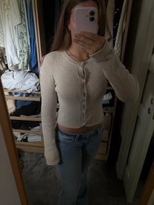 Beige ribbad kofta med knappar - Säljer en beige ribbad kofta med långa ärmar och knäppning framtill. Modellen är croppad och har en mjuk, stretchig passform som sitter snyggt på kroppen. Perfekt att styla med jeans eller kjol för en trendig look.