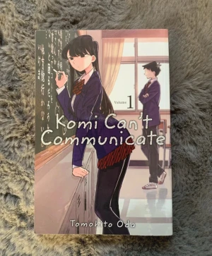 Komi Can't Communicate Volym 1 - Manga-bok på engelska av Tomohito Oda. Omslaget har en tecknad skolmiljö med karaktärer i skoluniform. Serien handlar om Shoko Komi och hennes utmaningar med social ångest och vänskap. Perfekt för dig som gillar slice of life och humoristisk skönlitteratur.