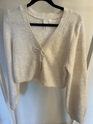 Gräddvit cropped stickad tröja med pärldetalj - Supersöt beige stickad tröja i croppad modell med djup v-ringning och en rosett av pärlor framtill. Tröjan är använd men i bra skick. Tröjan har långa ärmar och ribbad struktur, perfekt för lager på lager och trendiga outfits.