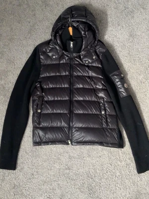 Svart pufferjacka från Moncler - Snygg svart pufferjacka från Moncler med quiltad kropp och stickade ärmar. Jackan har huva, dragkedja framtill, två fickor med tryckknappar och en Moncler-logga på ärmen. Perfekt för kalla vinterdagar och riktigt trendig look. Tar mer än gärna emot prisförslag då jag ska flytta och behöver pengar🤗