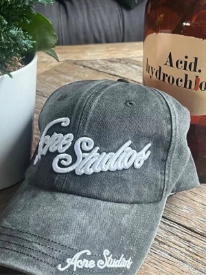 Grå keps från Acne Studios - Säljer en grå keps från Acne Studios med broderad vit logga framtill och på skärmen. Kepsen har klassisk böjd skärm och justerbar rem bak. Tillverkad i bomull för en skön och avslappnad look.