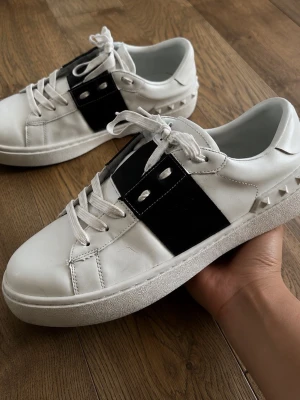 Vita sneakers med svart detalj - Snygga vita sneakers med en bred svart läderdetalj på sidan och coola nitar längs hälen. Skorna har vita snören och en klassisk låg siluett. Perfekta för dig som gillar en stilren men edgy look.