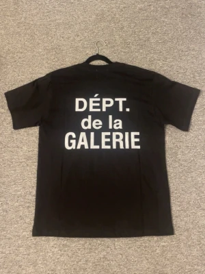 Svart t-shirt med vit text - Snygg svart t-shirt med stor vit text 'DÉPT. de la GALERIE' på ryggen. Klassisk rak passform och korta ärmar. Tillverkad i mjukt bomullstyg som känns skönt mot huden. Perfekt för dig som gillar streetwear och tydliga prints.
