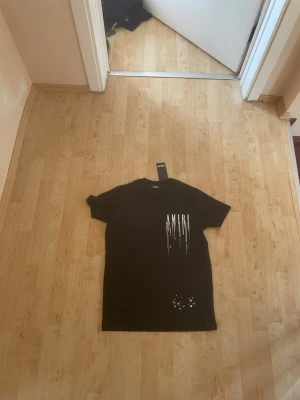 Svart AMIRI t-shirt med tryck - Snygg svart t-shirt från AMIRI med vitt tryck och färgstänk på framsidan. T-shirten har korta ärmar och en relaxed passform. Materialet känns mjukt och skönt, perfekt till streetwear-looken.