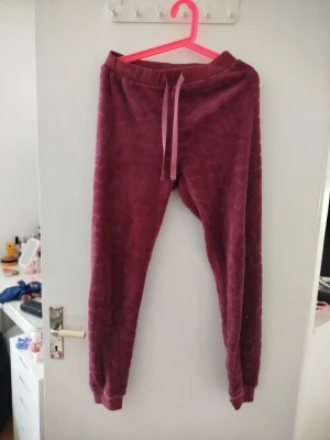 Vinröda mjukisbyxor med hjärtmönster - Supersköna vinröda mjukisbyxor i fleece som gör dom väldigt varma och mysiga med präglade hjärtan. Byxorna har resår i midjan och dragsko för bästa passform samt muddar vid bensluten. Perfekta för chill hemma eller mysiga kvällar.