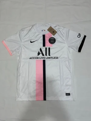 Vit PSG matchtröja med rosa och svart - Säljer en snygg vit PSG matchtröja från Nike med vertikala rosa och svarta ränder framtill. Tröjan har korta ärmar, klubbmärke på bröstet och sponsorlogga. Materialet är lätt och andas, perfekt för fotboll eller träning.