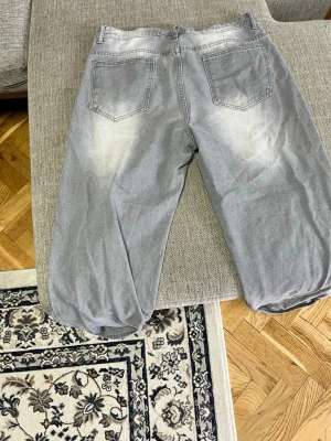 Gråa loose fit jeans med tvättade detaljer - Säljer ett par gråa loose fit jeans med tvättade och slitna detaljer framtill och baktill. Jeansen har fem fickor, raka ben och klassisk knappgylf. Perfekta för en avslappnad och trendig look.