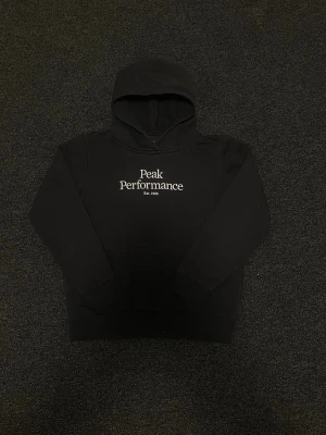 Svart hoodie från Peak Performance - Snygg svart hoodie från Peak Performance med vit broderad logga och texten 'Est. 1986' på bröstet. Klassisk modell med huva och känguruficka framtill. Perfekt för en avslappnad och sportig stil. ‼️pris kan diskuteras‼️