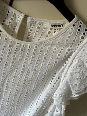 Vit broderad blus från Koton - Söt vit blus från Koton med broderade hålmönster och volangdetaljer på ärmarna. Blusen har rund halsringning och knapp i nacken. Tillverkad i ett lätt och luftigt tyg, perfekt för sommardagar.